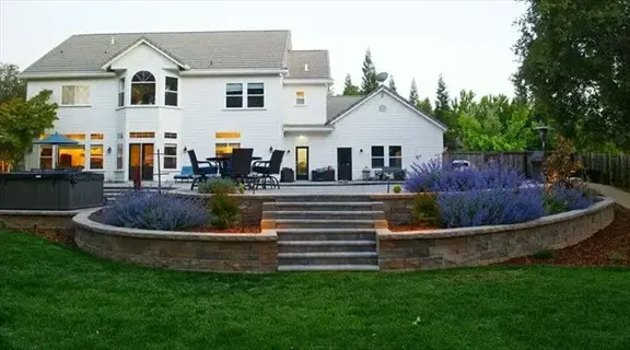 backyard featuring drought tolerant grass lawn, tiered stone patio with steps, and purple flowering shrubs under evening sky