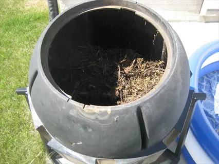 black rotating compost drum filled with organic debris, outdoors on a stand near grass and a building