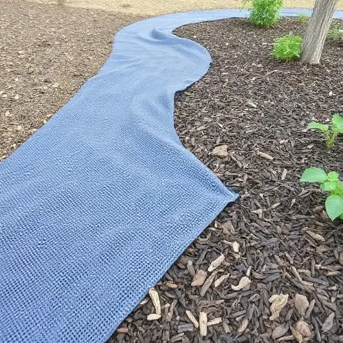 blue landscape fabric garden bed laid on mulch path with trees and plants in background