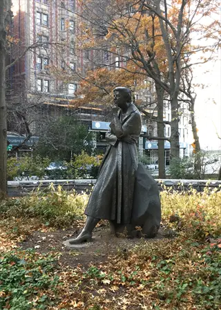 bronze statue of eleanor roosevelt seated on a rock in a park with autumn foliage and city buildings in the background