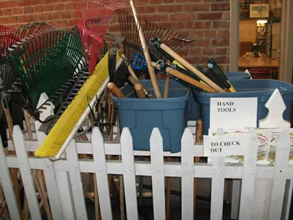 community tool library garden with shared gardening tools (rakes, brooms) in bins marked 'hand tools' and 'to check out' near white picket fence