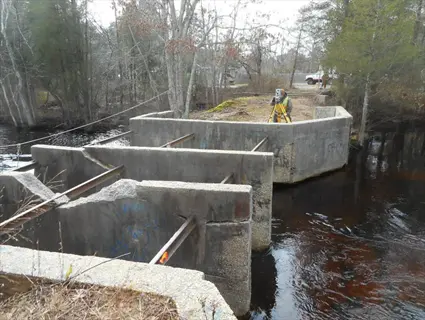 concrete aquatic fish barrier (weir/dam) with surveyor, graffiti, and flowing water in forested setting