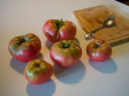 five ripe german johnson tomatoes with pink-red skin and green shoulders on a kitchen countertop, beside a wooden cutting board and spoon