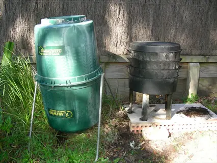 green barrel compost tumbler and black multi-tiered composter against wooden fence with grass