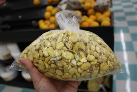 hand holding a clear freezer bag filled with fava bean seeds in a grocery store, background of oranges and produce