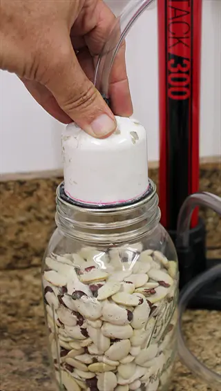 hand using vacuum sealer to create vacuum sealed seeds in a mason jar filled with dried beans, with 'tack 300' vacuum device visible