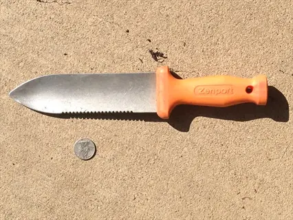 hori hori garden knife with orange handle and metal blade beside a coin on concrete