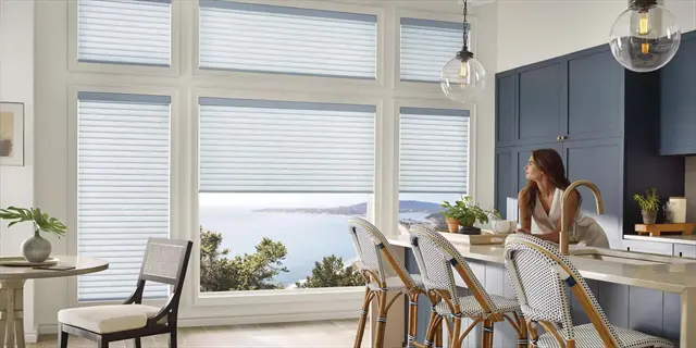 light diffusing blinds in a coastal kitchen with ocean view, woman standing at kitchen island with wicker chairs and pendant lights
