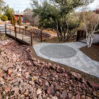low maintenance native landscape featuring rock garden, wooden bridge, paved walkway, and drought-tolerant native trees under a sunny sky
