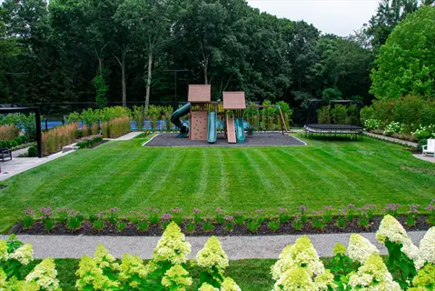 lush backyard lawn family play area featuring playground set, trampoline, and manicured garden beds with blooming flowers