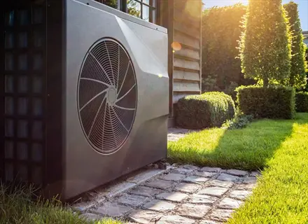 outdoor greenhouse heat pump unit in sunny garden setting with cobblestone path, trimmed hedges, and wooden building