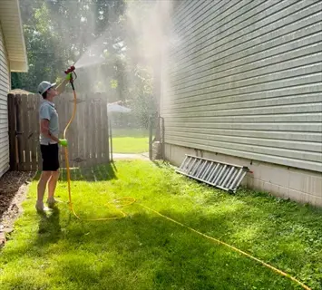 person using a house foundation watering hose in a sunny backyard with a ladder and fence