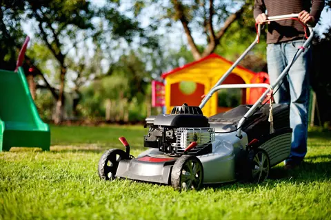 person using lawn mower cutting grass in backyard with colorful playground equipment