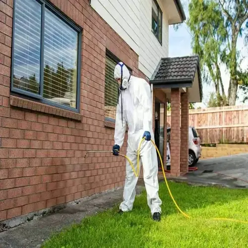 pest control technician in white protective suit spraying residential house exterior with handheld nozzle