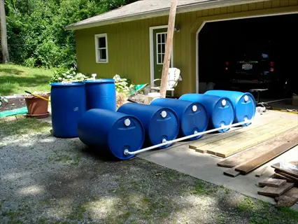 residential rain barrel system with interconnected blue barrels and white pipes in a driveway, part of a home rainwater harvesting setup