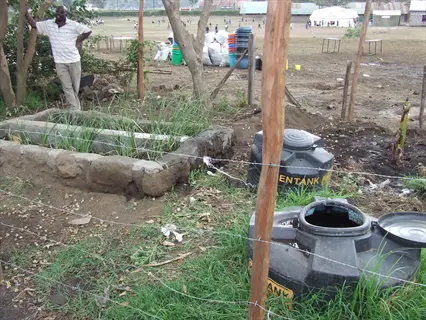 rural greywater filtration system featuring kentank storage tanks and a constructed wetland (stone-lined plant bed) in a fenced outdoor environment, with an observer nearby
