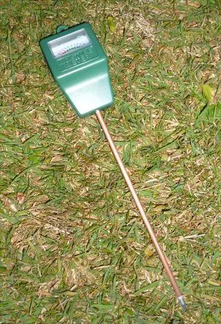 soil moisture meter: green handheld device with analog gauge and metal probe resting on grassy surface, designed for monitoring soil moisture levels