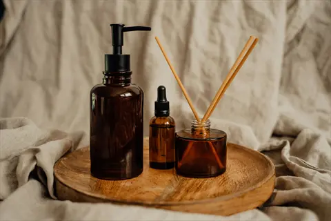 three amber glass bottles (pump dispenser, dropper, and reed diffuser) on wooden tray with linen background