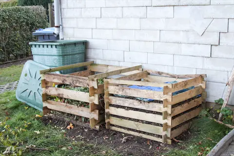 two diy pallet compost bins against a building wall with green compost bin and lid nearby