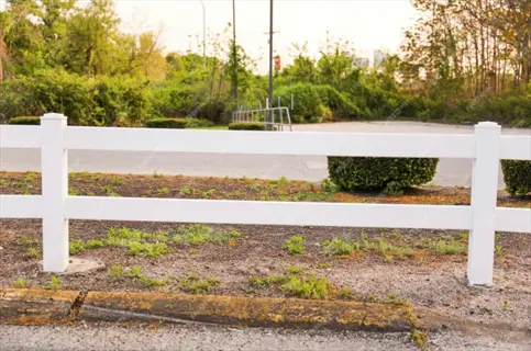 white split rail garden fence bordering a landscaped area with shrubs, trees, and a paved road in the background