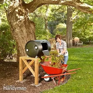woman adding garden waste to large compost tumbler on wooden platform with wheelbarrow in backyard