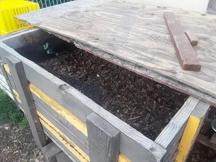 wooden insulated compost tumbler with yellow and gray slats, partially open to reveal soil and organic matter inside