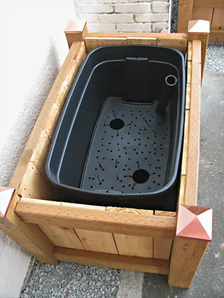 wooden self watering planter with black plastic reservoir liner and water level tube against light-colored wall