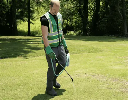 worker in green safety vest applying mulch for weed prevention on a sunny lawn with trees in background